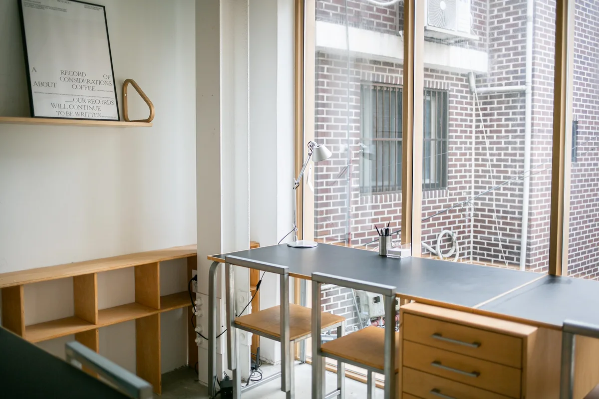 Window-side desks, wooden chairs, a task lamp and a framed coffee poster inside Protokoll’s Sangsu cafe.